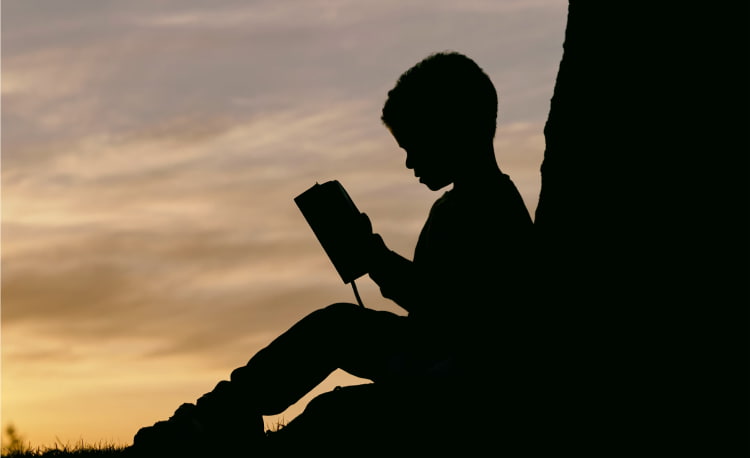 Sillouhette of a young boy leaning on a tree reading a book